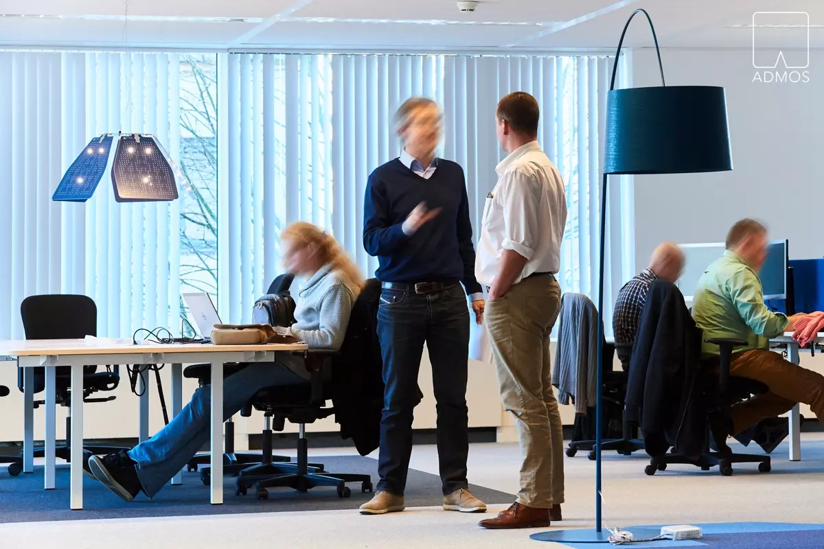 Modern kantoor met werkende en overleggende mensen, blauwe designlampen en witte bureaus.